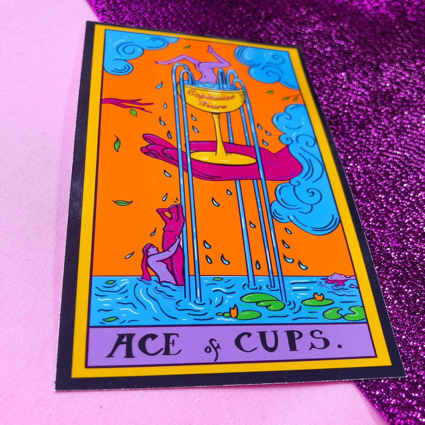 Tarot card sticker based on the Ace Of Cups. Original artwork, a pink hand holds a gold wine glass with water flowing out into the river below where there are naked people dancing in the river. The cup reads Capitalist Tears.