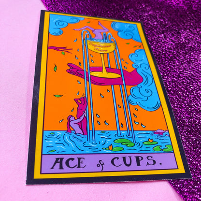 Tarot card sticker based on the Ace Of Cups. Original artwork, a pink hand holds a gold wine glass with water flowing out into the river below where there are naked people dancing in the river. The cup reads Capitalist Tears.